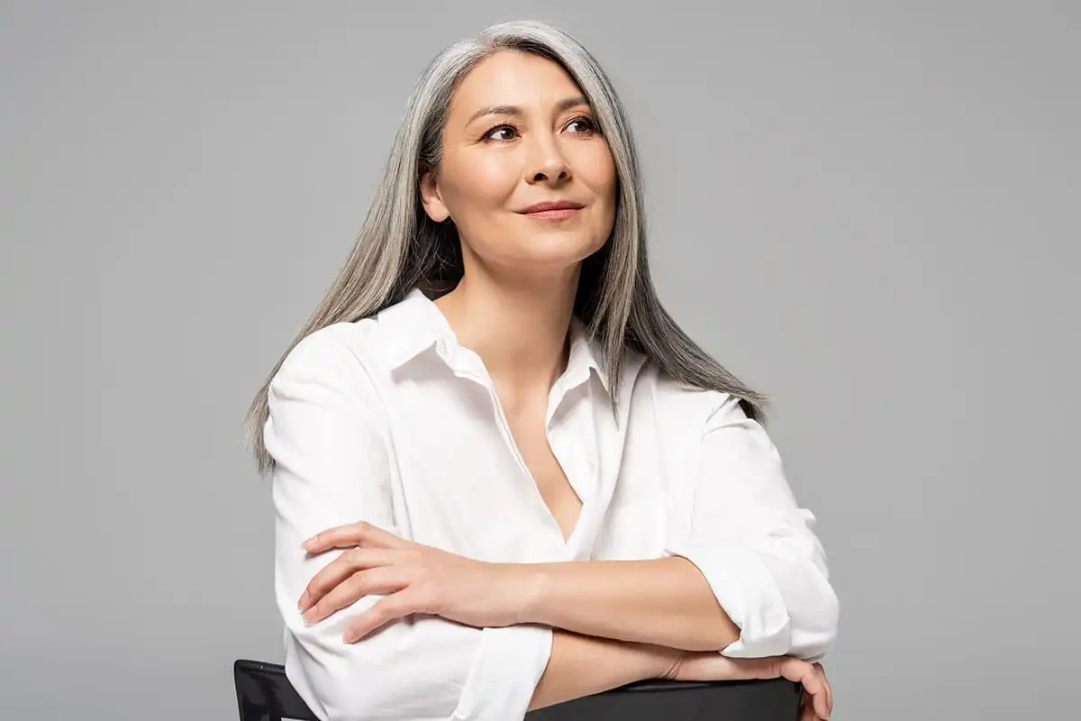Woman with gray hair wearing white shirt, arms crossed, looking upward thoughtfully