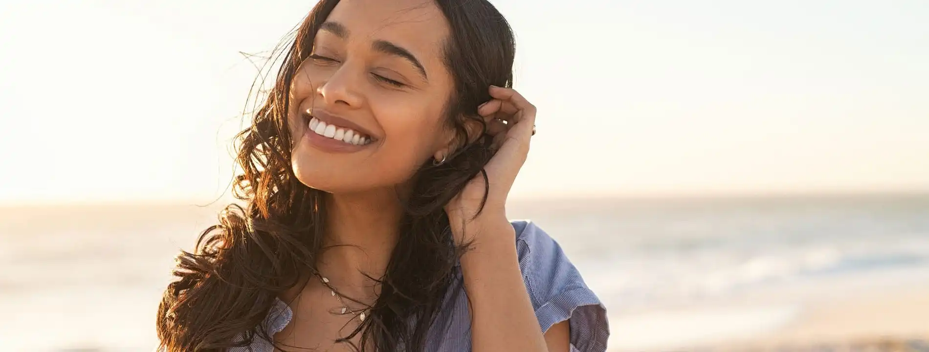 Woman smiling at beach at sunset while touching her hair, wearing blue shirt