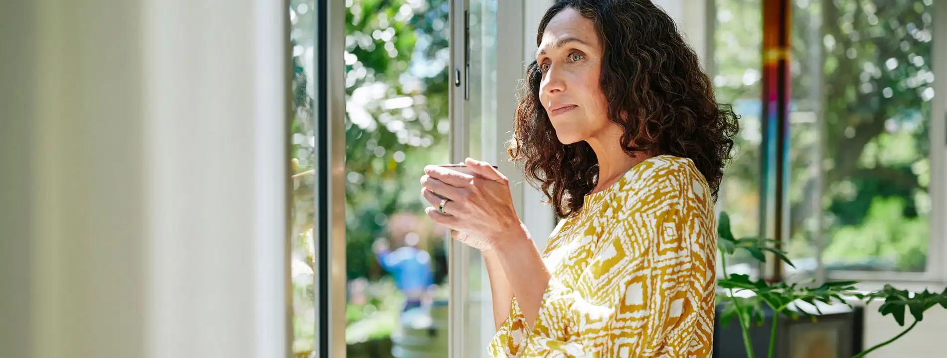 Woman holding a beverage near large windows overlooking a garden, wearing yellow geometric patterned top