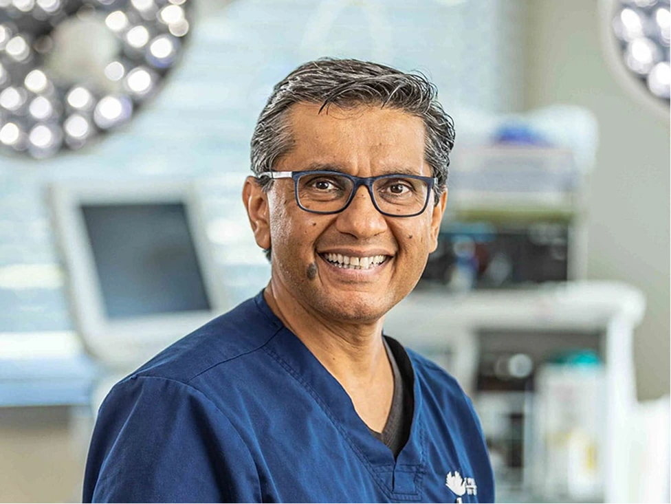 Plastic surgeon in blue scrubs and glasses smiling in surgical suite with overhead lights and medical equipment