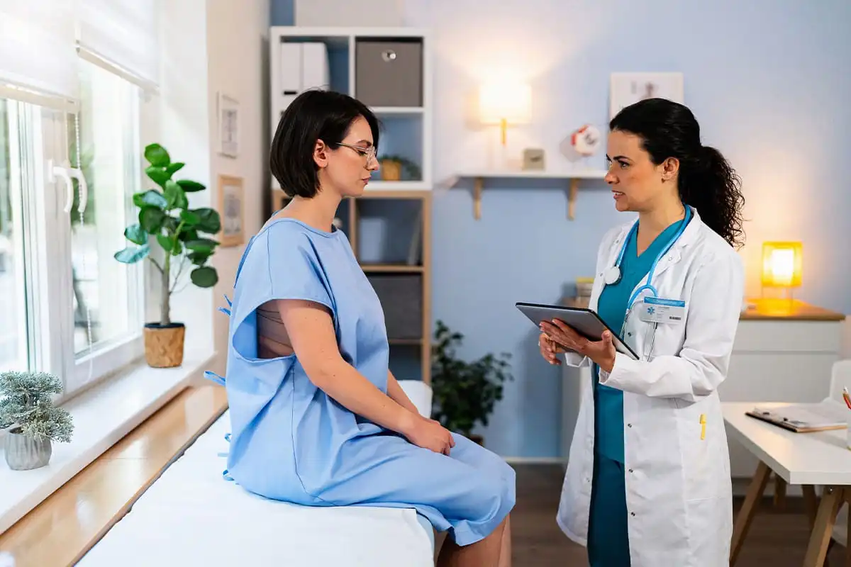Plastic surgeon in white coat and teal scrubs consulting with patient in blue hospital gown while reviewing tablet in bright clinical office
