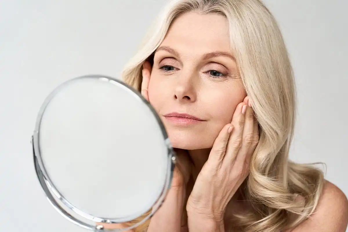 Blonde woman examining her face in handheld mirror with hand on cheek