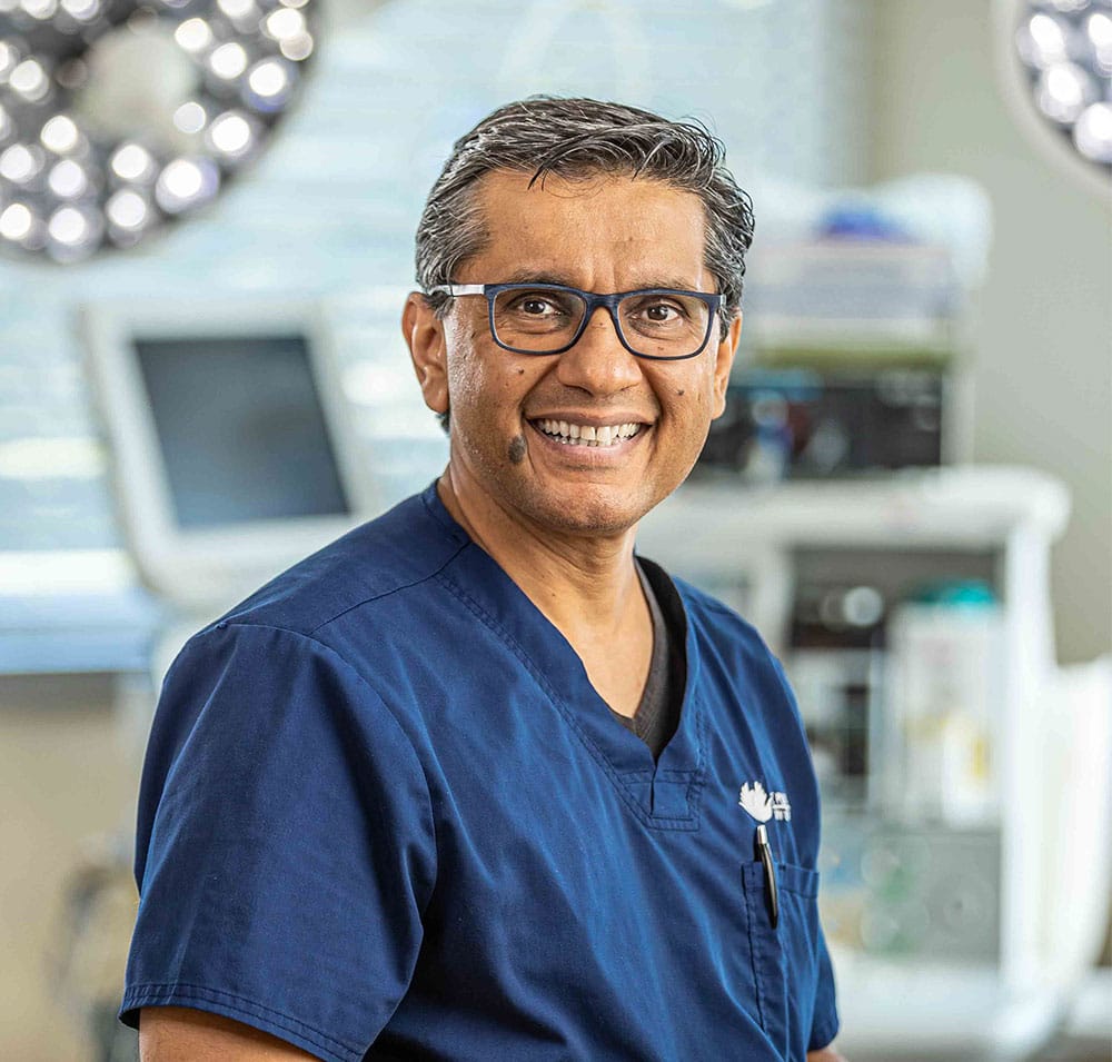 Surgeon in blue scrubs and glasses smiling in operating room with surgical lights and medical equipment