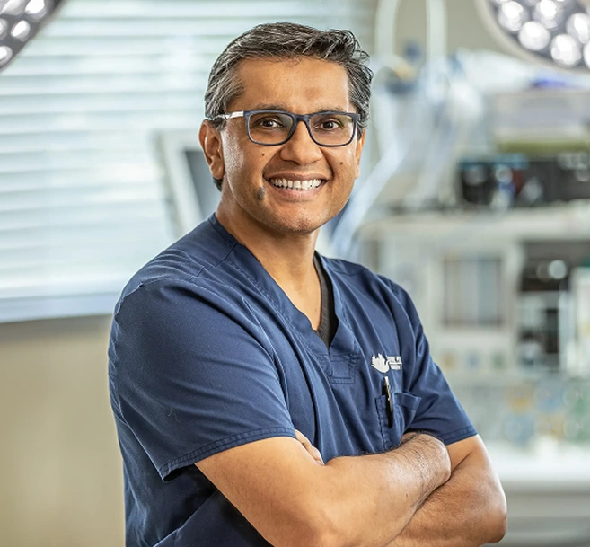 Plastic surgeon in blue scrubs and glasses smiling with arms crossed in surgical suite