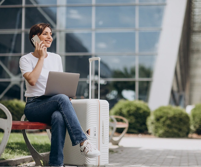 Woman on outdoor bench talking on phone while holding laptop and sitting next to white luggage