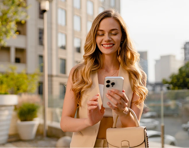 Woman on rooftop holding smartphone and smiling, wearing beige blazer and shoulder bag with city buildings in background