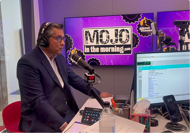 Man in dark suit wearing headphones speaking into microphone at radio station studio with MOJO in the morning logo on screen
