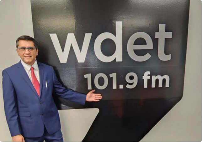 Man in blue suit with pink tie standing next to WDET 101.9 FM radio station sign