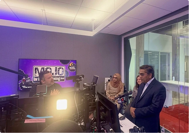 Radio host interviewing woman in hijab and man in suit at studio desk with camera and purple lighting