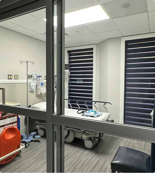 Operating room view through glass partition showing surgical bed and overhead lights